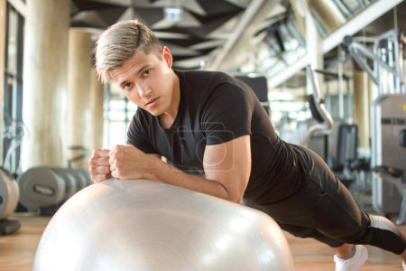 Téléchargez les photos : Beau jeune homme faisant des exercices de planche sur balle de fitness à la salle de gym - en image libre de droit
