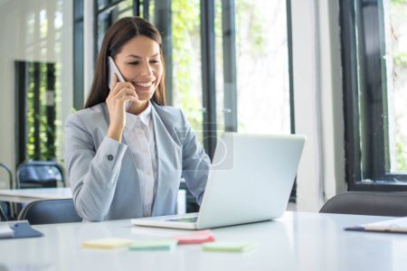 Foto de Mujer de negocios hablando por teléfono mientras trabaja en el ordenador portátil en la oficina. Concepto empresarial. - Imagen libre de derechos
