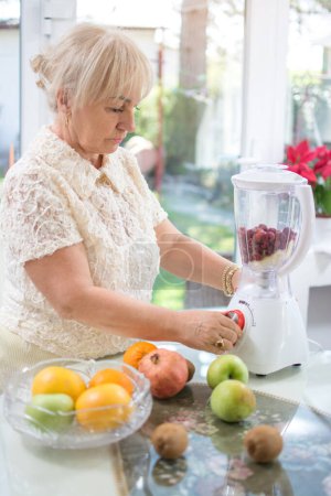 Foto de Hermosa mujer mayor preparando batido orgánico vegano, utilizando licuadora en la cocina. - Imagen libre de derechos