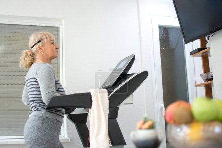 Photo for Active senior woman in sportswear walking on treadmill and watching tv at home. Home quarantine, Corona virus, active seniors concept. - Royalty Free Image