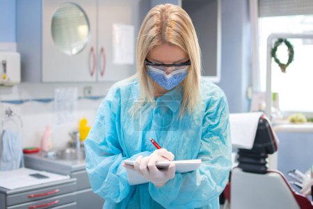 Photo for Portrait of female dentist in a protective suit, shield, mask and eyeglasses writing notes to planner book in dental office. - Royalty Free Image