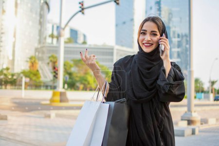 Foto de Mujer árabe en Abaya hablando por teléfono celular y sosteniendo bolsas en la calle - Imagen libre de derechos