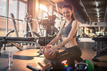 Téléchargez les photos : Fille sportive tenant ballon de remise en forme et de détente tout en étant assis sur tapis d'exercice pendant la pause d'entraînement dans la salle de gym. - en image libre de droit