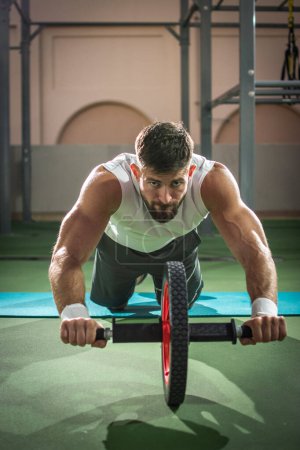 Photo for Handsome sportsman doing ab wheel rollout exercise and looking forward while working out at gym. - Royalty Free Image