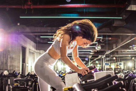 Photo for Side shot of attractive young brunette sportswoman riding exercise bike during cycling workout in gym - Royalty Free Image