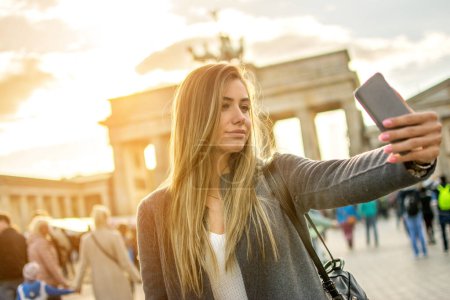 Téléchargez les photos : Belle jeune femme prenant selfie photo devant Brandenburger Tor à Berlin au coucher du soleil. - en image libre de droit