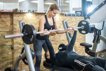 Photo for Handsome young man lifting weights with assistance of attractive girl in health club. - Royalty Free Image