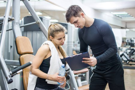 Photo for Young sporty woman and her personal trainer looking together at sports results on clipboard in gym. - Royalty Free Image