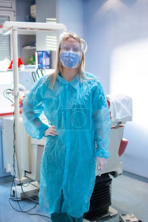 Foto de Dentista femenina con traje protector con escudo protector y máscara de pie en el consultorio dental - Imagen libre de derechos