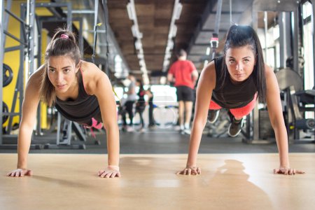 Photo for Two sporty girls doing push ups while their legs are on suspension strap at gym. - Royalty Free Image