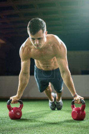 Photo for Concentrated muscular young man in plank position workout with kettle bells equipment in the gym. - Royalty Free Image