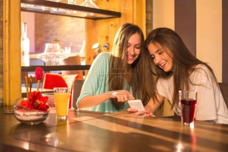 Photo for Two young teenage girl using phone while having a drink together in bar - Royalty Free Image