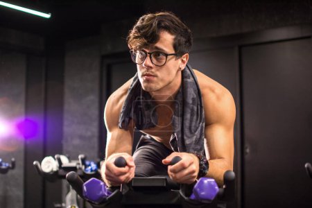 Téléchargez les photos : Jeune homme beau torse nu avec des lunettes équitation vélo d'exercice dans la salle de gym - en image libre de droit