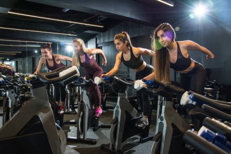 Foto de Grupo de chicas de fitness montando bicicletas estáticas juntas en clase de ciclismo en el gimnasio. - Imagen libre de derechos