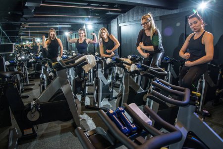 Photo for Strong sporty women showing and flexing their muscles while sitting on exercise bikes in gym - Royalty Free Image