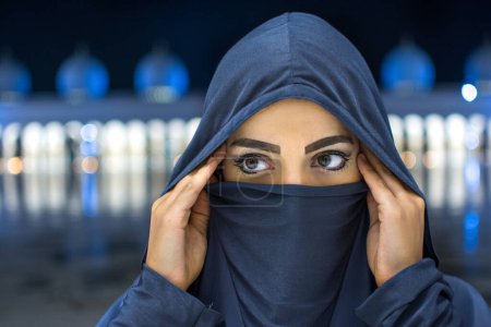 Téléchargez les photos : Gros plan portrait de belle jeune femme musulmane portant le hijab regardant loin avec la mosquée à l'arrière-plan pendant la nuit. - en image libre de droit