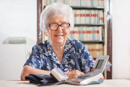 Foto de Feliz anciana sentada cerca de la mesa con monitores de presión arterial - Imagen libre de derechos