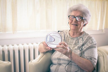 Glückliche Großmutter hört Musik auf CD-Player, während sie zu Hause im Sessel sitzt.