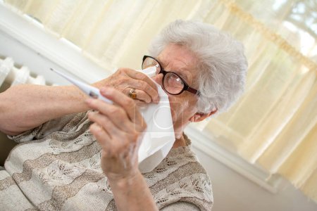 Foto de Mujer mayor comprobando la temperatura corporal con termómetro y limpiando la nariz con un papel tisú. - Imagen libre de derechos