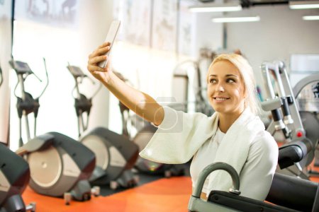 Téléchargez les photos : Jeune fille blonde faisant selfie photo sur smartphone pendant la pause d'entraînement dans la salle de gym. - en image libre de droit