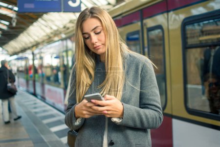Foto de Mujer joven usando el teléfono móvil mientras espera el tren en la estación de tren. - Imagen libre de derechos
