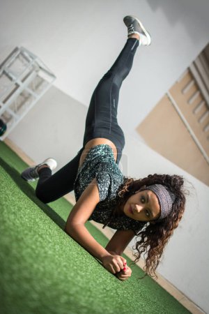Mujer joven en ropa deportiva haciendo ejercicio de patada de burro, mientras se arrodilla en el suelo y levanta una pierna al aire libre.