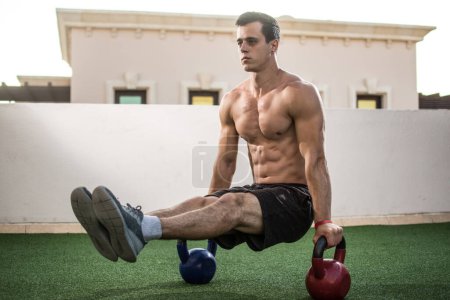 Muscular shirtless man workout with kettlebells in L Sit position outdoors.