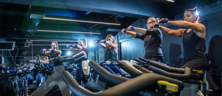 Foto de Vista panorámica de las chicas deportivas haciendo ejercicios con pesas mientras monta bicicleta de fitness en el gimnasio. - Imagen libre de derechos