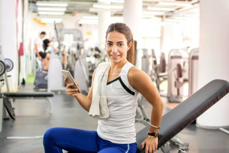 Téléchargez les photos : Jeune femme sportive avec smartphone assis sur une machine de gym dans un centre de remise en forme. - en image libre de droit
