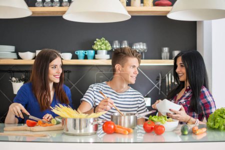 Téléchargez les photos : Trois jeunes amis cuisinent et s'amusent dans la cuisine. Ils préparent la nourriture ensemble et rient - en image libre de droit