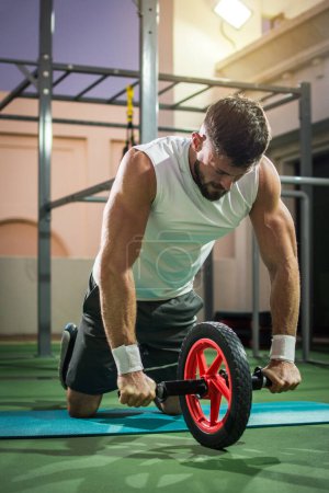 Muscular man doing exercises with abdominal toning wheel at gym.