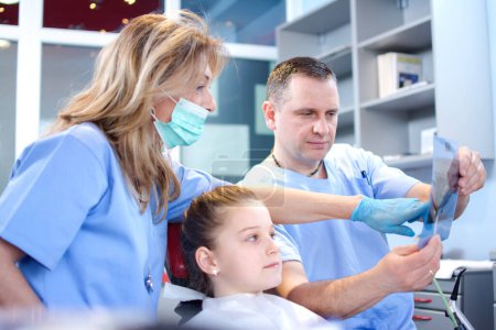Téléchargez les photos : Dentiste professionnelle avec l'aide d'un médecin masculin examinant une image radiographique d'une petite fille patiente dans une clinique dentaire. - en image libre de droit