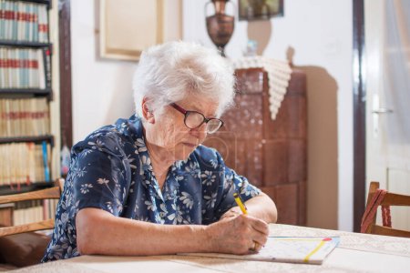Photo for Elderly woman with eyeglasses sitting near the table and doing crossword - Royalty Free Image