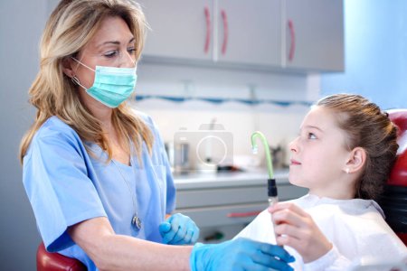 Téléchargez les photos : Fille patiente tenant un tube d'aspiration près d'une dentiste professionnelle dans un cabinet dentaire - en image libre de droit