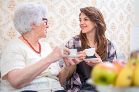 Téléchargez les photos : Portrait d'une grand-mère et de sa petite-fille buvant du café et parlant à la maison - en image libre de droit