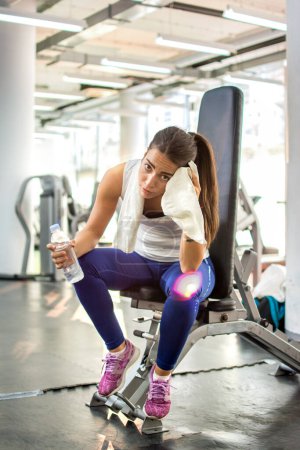Téléchargez les photos : Femme sportive fatiguée avec serviette et bouteille d'eau assise sur une machine de gym dans un centre de remise en forme. - en image libre de droit