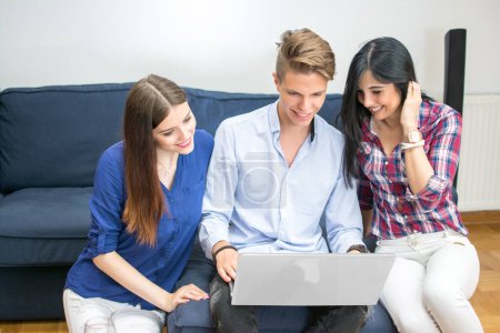 Téléchargez les photos : Trois jeunes amis utilisant un ordinateur portable assis sur le canapé à la maison - en image libre de droit
