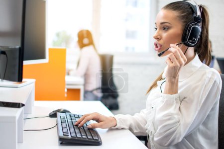 Photo for IT service desk female operator talking to client using headset and working on computer in call center office. - Royalty Free Image