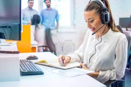 Foto de Operadora femenina con auriculares escribiendo notas mientras habla con el cliente en el centro de atención al cliente. - Imagen libre de derechos