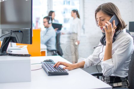 Mujer preocupada en ropa de negocios hablando por teléfono móvil, recibiendo malas noticias y sintiéndose deprimida mientras trabaja en la computadora en la oficina.