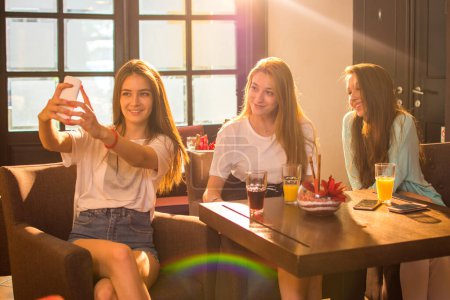 Photo for Group of three teenage girls taking a selfie photo for social media while sitting together in a cafe - Royalty Free Image
