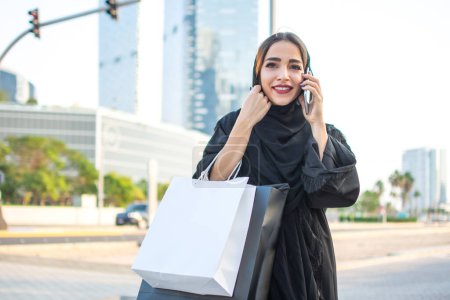 Photo for Beautiful Arabic woman carrying shopping bags and talking on mobile phone on a city street - Royalty Free Image