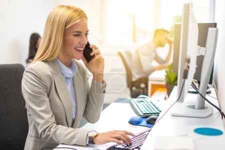 Téléchargez les photos : Jeune femme blonde parlant sur le téléphone portable et souriant tout en étant assise à son lieu de travail au bureau et en regardant l'écran d'ordinateur - en image libre de droit