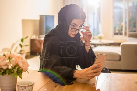 Photo for Beautiful young Arab woman with eyeglasses using smart phone at home. - Royalty Free Image