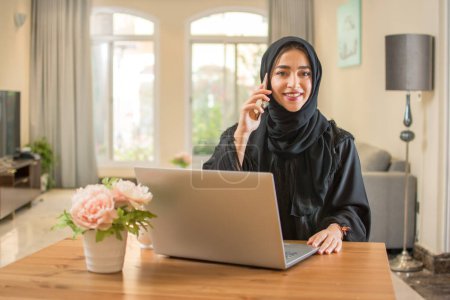 Photo for Beautiful young Arab woman wearing abaya talking on mobile phone and using laptop at home - Royalty Free Image