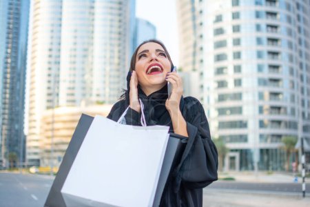 Téléchargez les photos : Joyeux jeune femme arabe en vêtements Abaya avec des sacs à provisions rire tout en parlant avec un ami sur téléphone portable dans la rue de la ville. - en image libre de droit