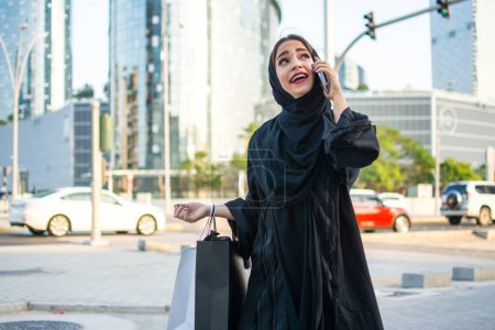 Téléchargez les photos : Belle femme arabe portant des sacs à provisions et parlant sur un téléphone portable dans la rue de la ville - en image libre de droit