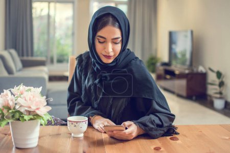 Photo for Beautiful young muslim woman in hijab using smart phone during morning coffee break at home. - Royalty Free Image