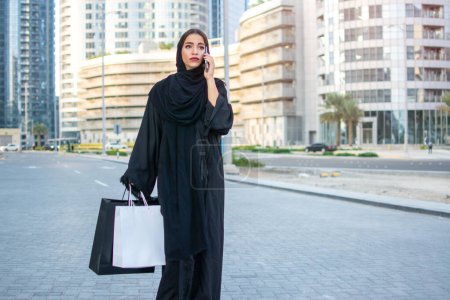 Téléchargez les photos : Belle arabe du Moyen-Orient portant Abaya, marchant dans la rue de la ville avec des sacs à provisions et parlant sur son téléphone intelligent. - en image libre de droit