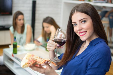 Téléchargez les photos : Jeune femme souriante tenant une tranche de pizza et un verre de vin rouge assis avec ses amis à la maison - en image libre de droit